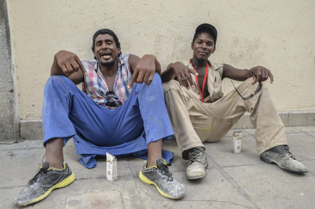 Dos hombres constructores toman ron sentados en la acera de un barrio habanero. Con las piernas abiertas en pose de macho y de hombría. Una forma de ser hombres en Cuba y mostrar una cultura patriarcal.