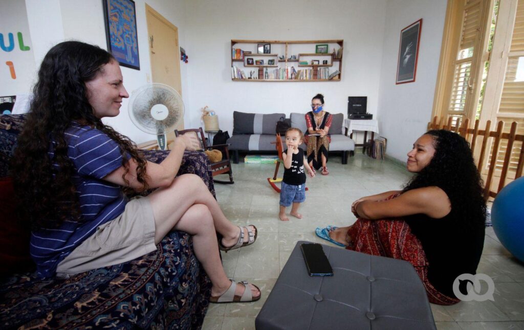 La familia formada por Dachelys y Hope en la sala de su casa, en su vida cotidiana. La pareja de mujeres se mira con satisfacción. El niño juega. Es el primer bebé con dos madres reconocido en Cuba. (Madre)