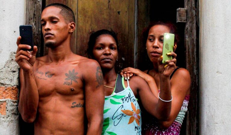 Dos mujeres y hombre consultan sus teléfonos celulares en Cuba. Están en el umbral de su casa. Matria